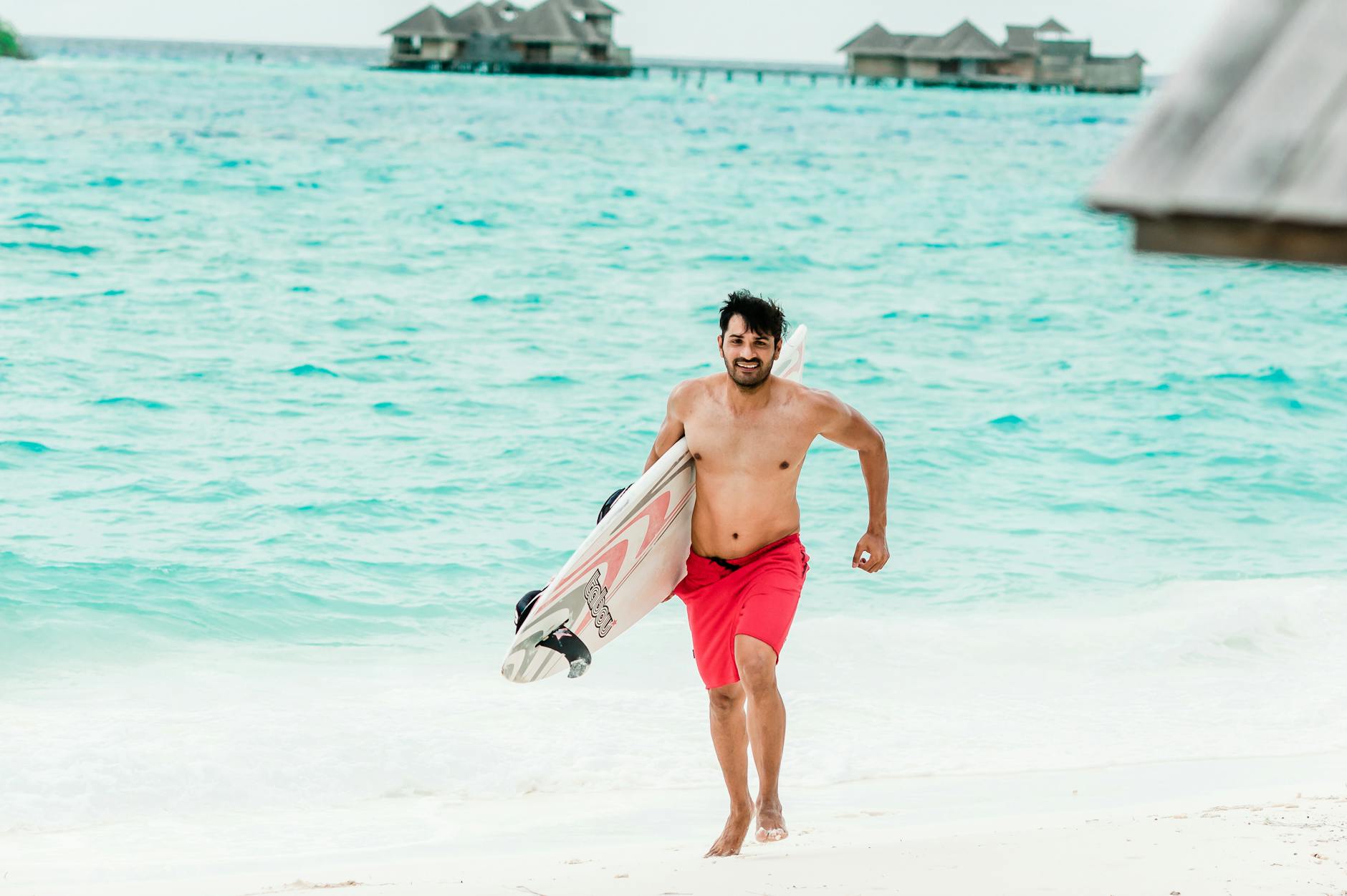 A man in red shorts joyfully runs on a Maldives beach carrying a surfboard.