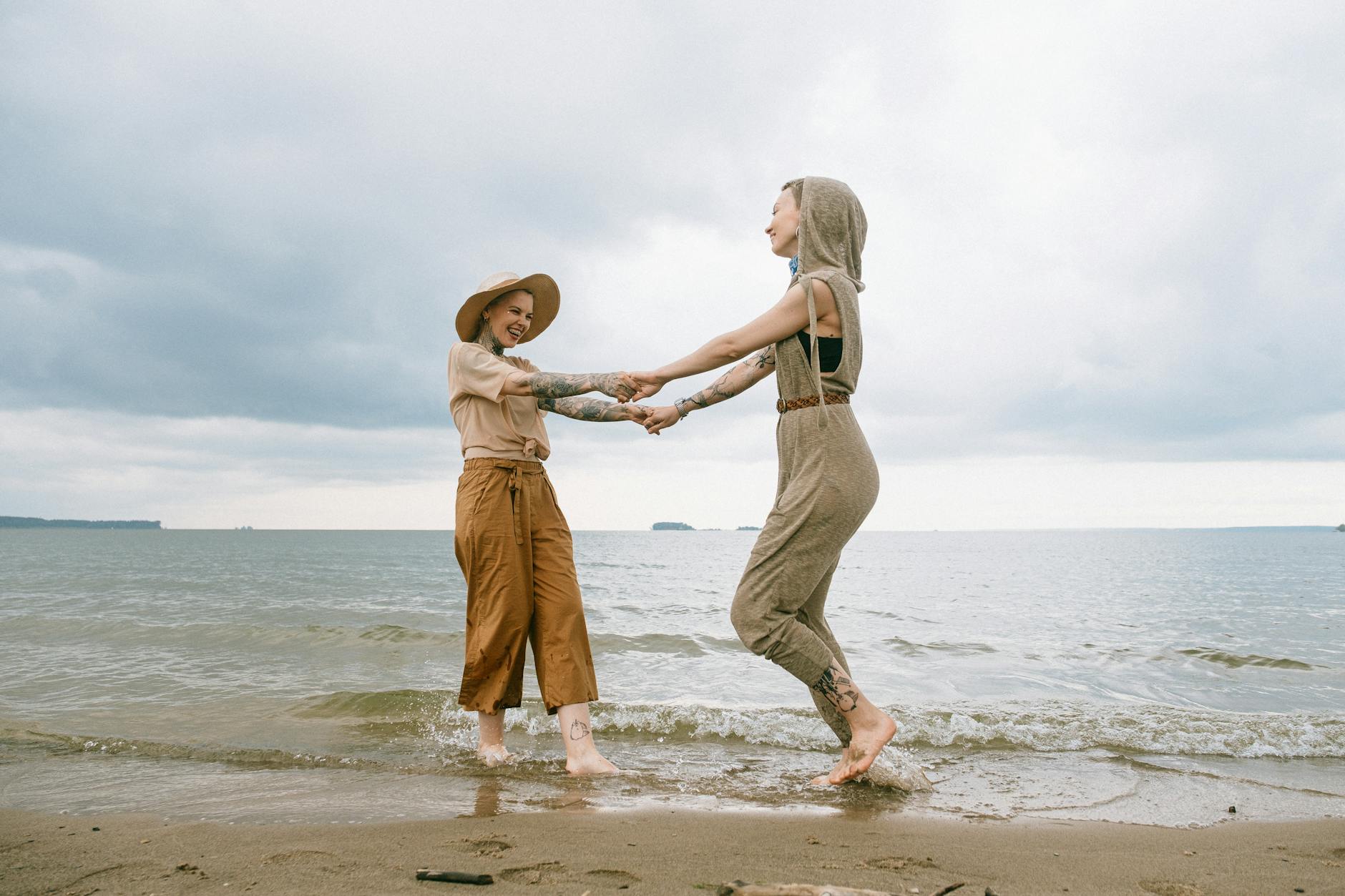 Two friends holding hands and dancing joyfully on a sandy beach by the sea, embodying happiness and freedom.
