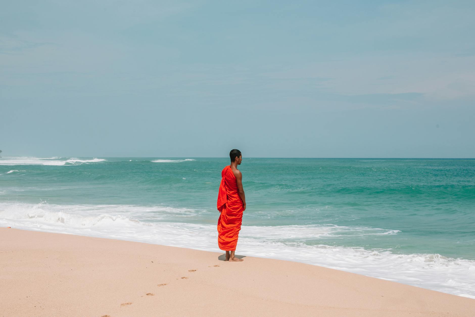 A Buddhist monk in orange robe meditates peacefully by the ocean shore, capturing tranquility.