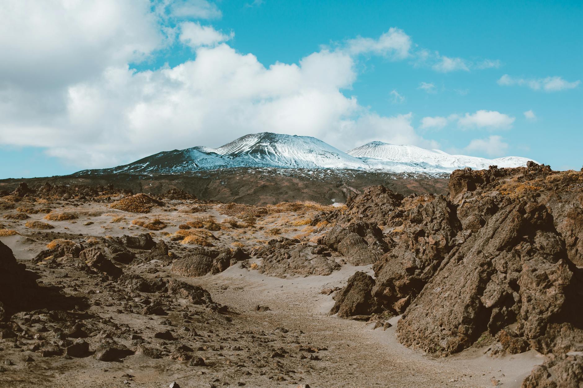 Picturesque view of distant mountains with snow on slopes surrounded by rocky terrain under cloud sky