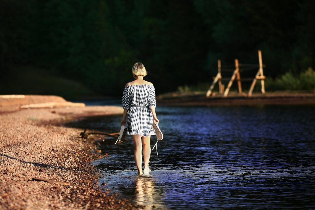 Home A woman in a dress walking along a riverbank, holding shoes in hand.