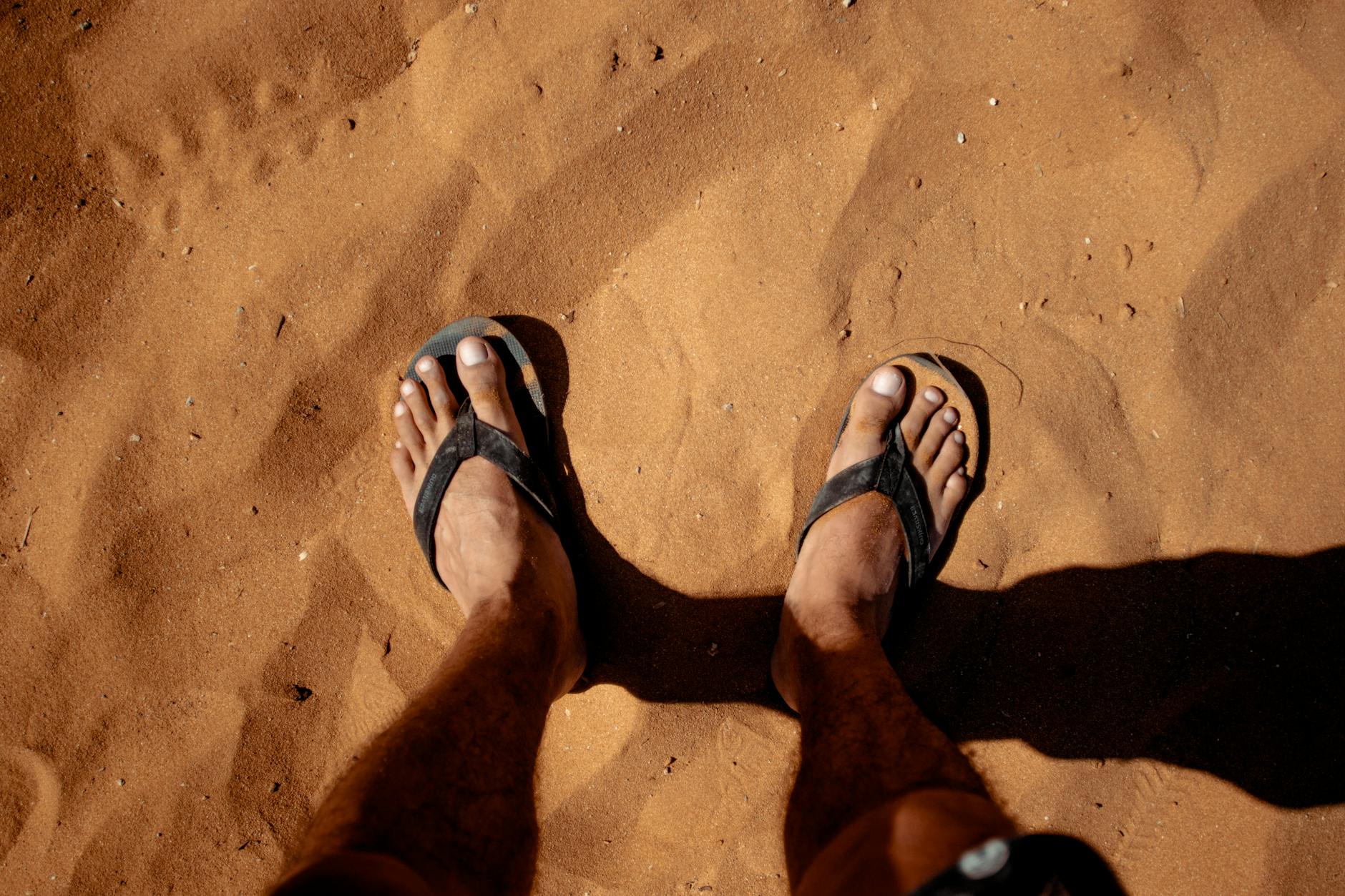 Close-up of feet in flip flops standing on sunlit sandy ground, casting shadows.