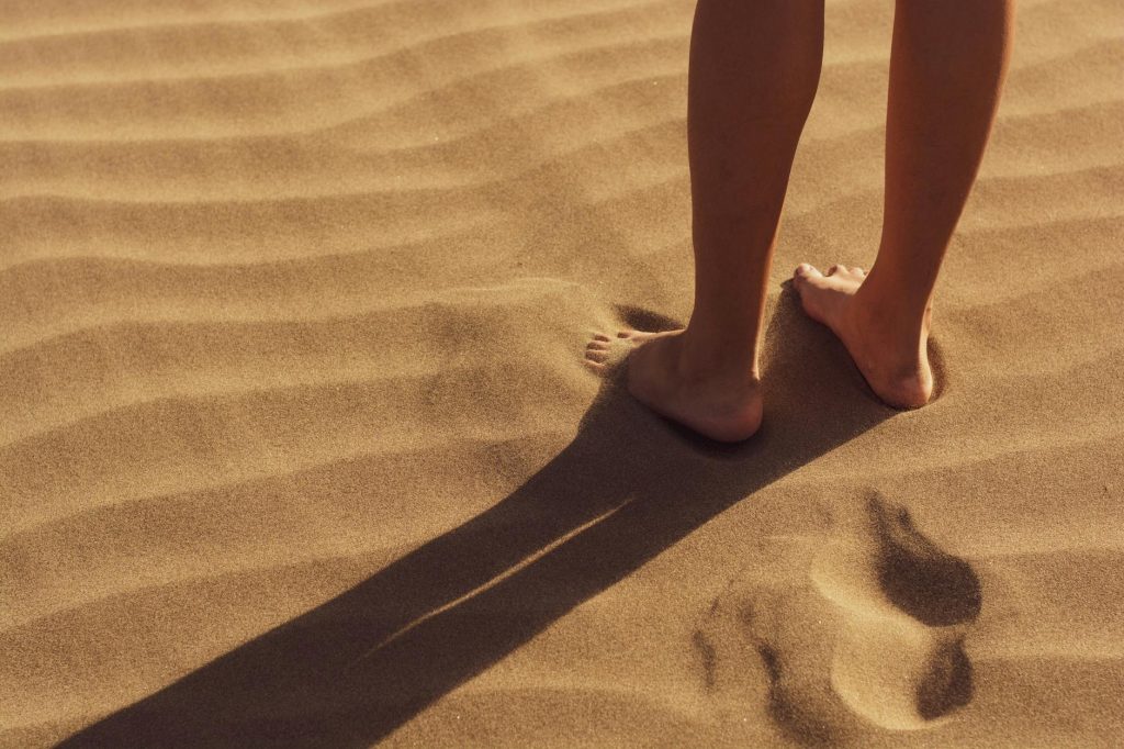 Home Close-up of a person standing barefoot on sunlit sand with visible footprints.