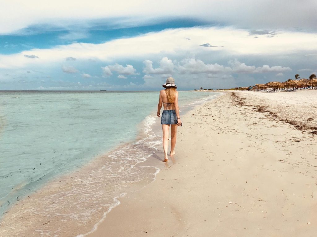 Home A woman walks along a serene tropical beach, enjoying a peaceful summer day.